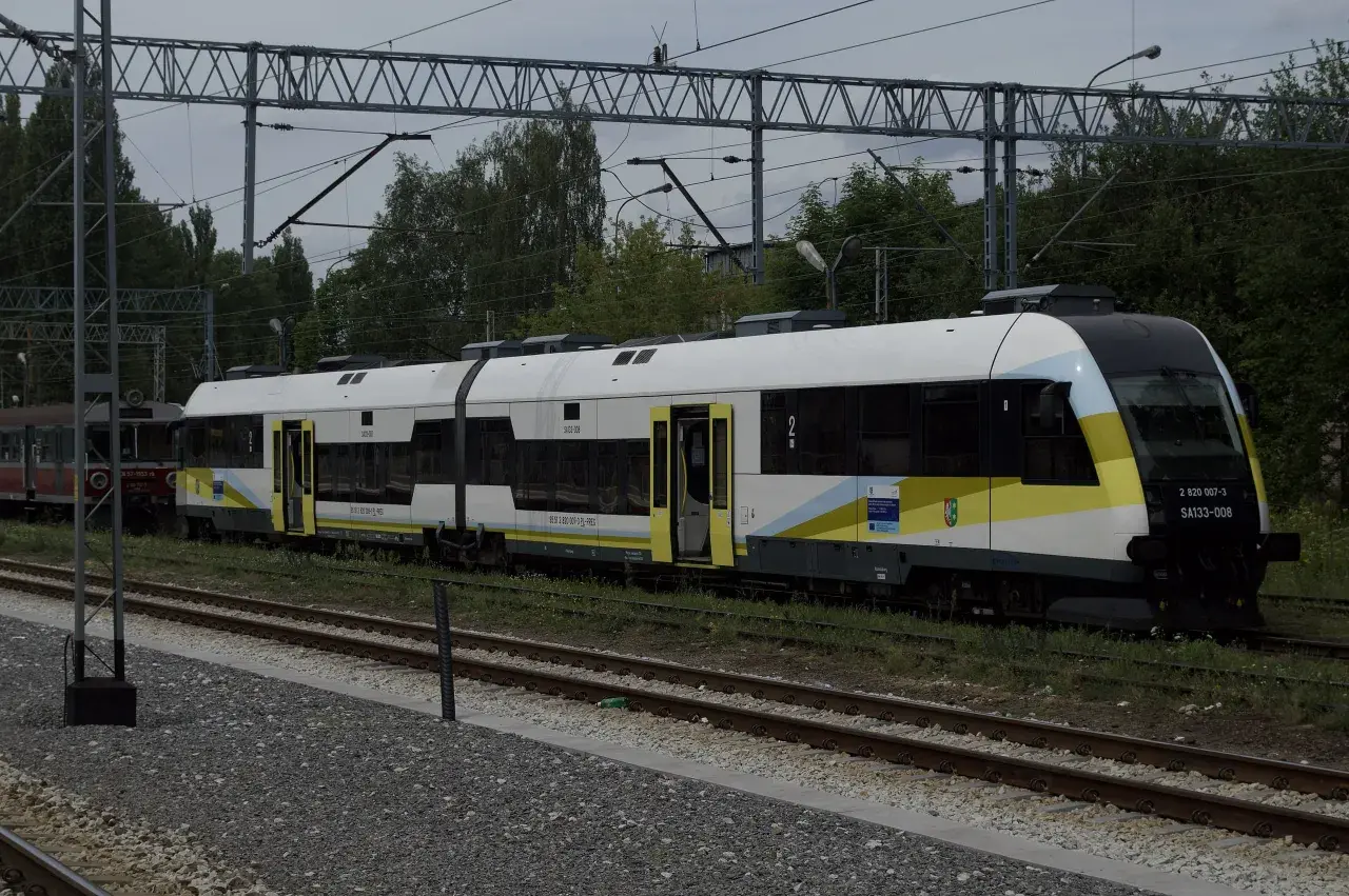Nowy pociąg SA133-008, typu Pesa Elf, z charakterystycznym malowaniem w barwach żółto-biało-niebieskich, stoi na torach. Obok widoczny starszy wagon.