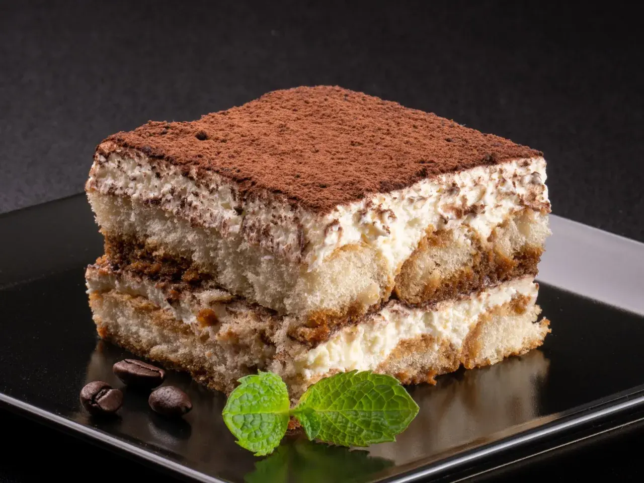 Kawałek tiramisu posypany kakao, z listkami mięty i ziarnami kawy na czarnym talerzu.