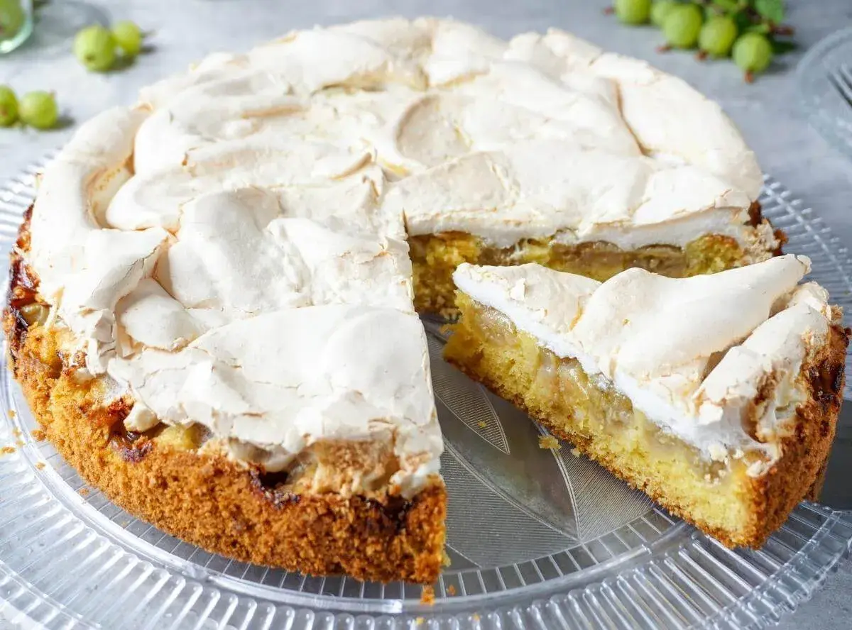 Ein saftiger Stachelbeer Baiser Kuchen mit goldbraunem Boden und fluffiger Baiserhaube, ein Stück ist angeschnitten.