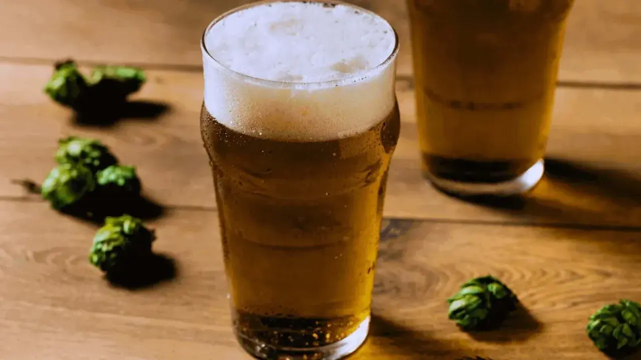 Un bicchiere di birra dorata con schiuma abbondante, circondato da luppoli verdi su un tavolo di legno. Perfetto per una festa della birra!