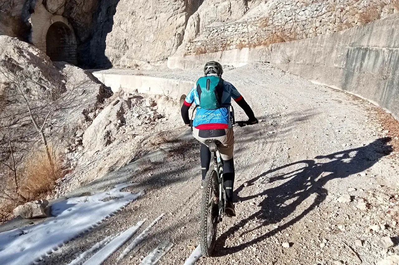 Ciclista in mountain bike su sentiero sterrato, con neve residua e un tunnel scavato nella roccia. Un'avventura tra le **Tignale cascate**.