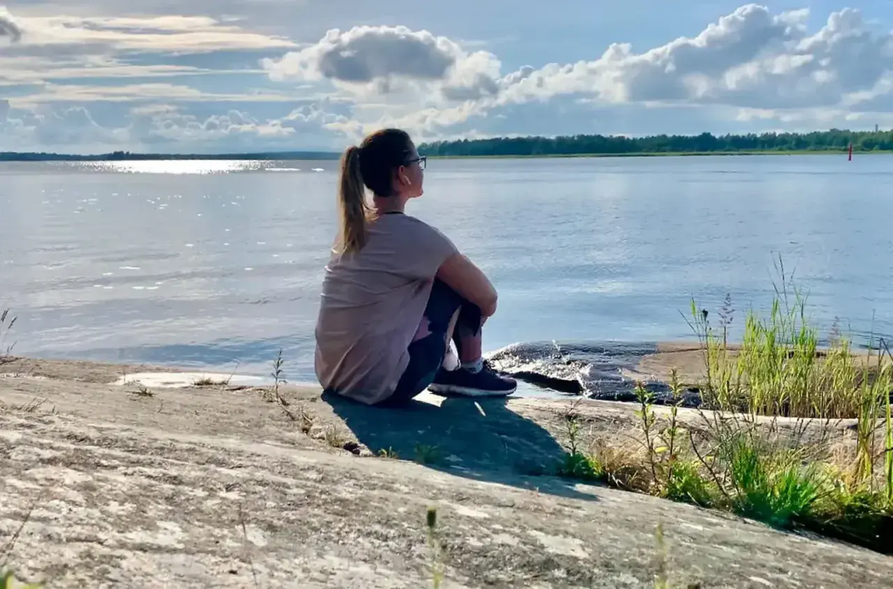 Kvinna sitter på en klippa vid havet, övar mindfulness för att minska stress. Solen glittrar på vattnet.