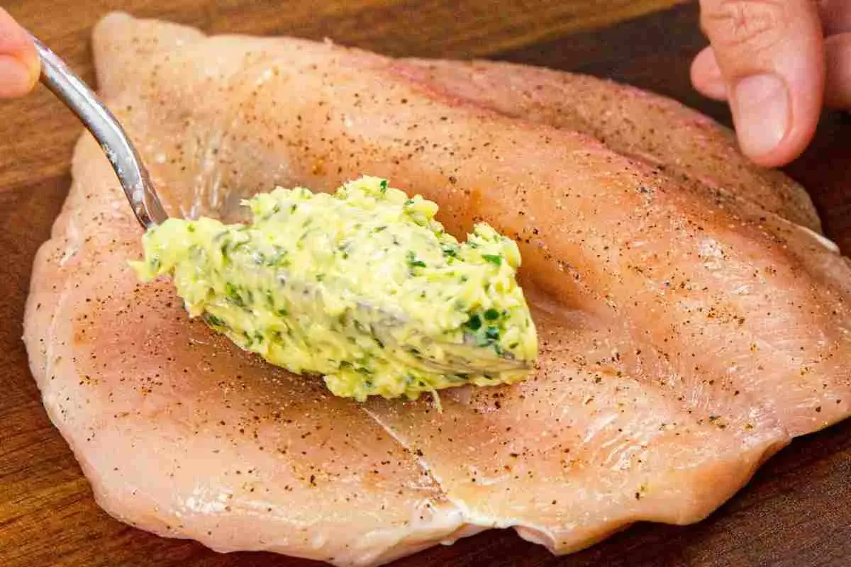 Un cucchiaio di burro alle erbe viene spalmato su un petto di pollo crudo, pronto per essere cucinato.