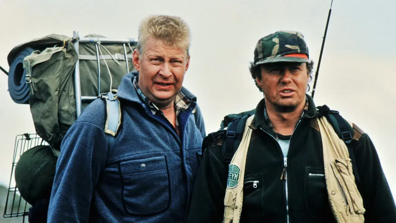 Two men in outdoor gear, one with a large backpack, stand against a pale sky.