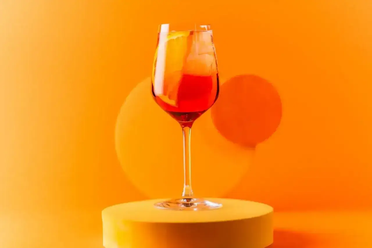 Orzeźwiający spritz campari z plasterkiem pomarańczy i lodem, prezentowany na żółtym podeście na tle pomarańczowego tła.