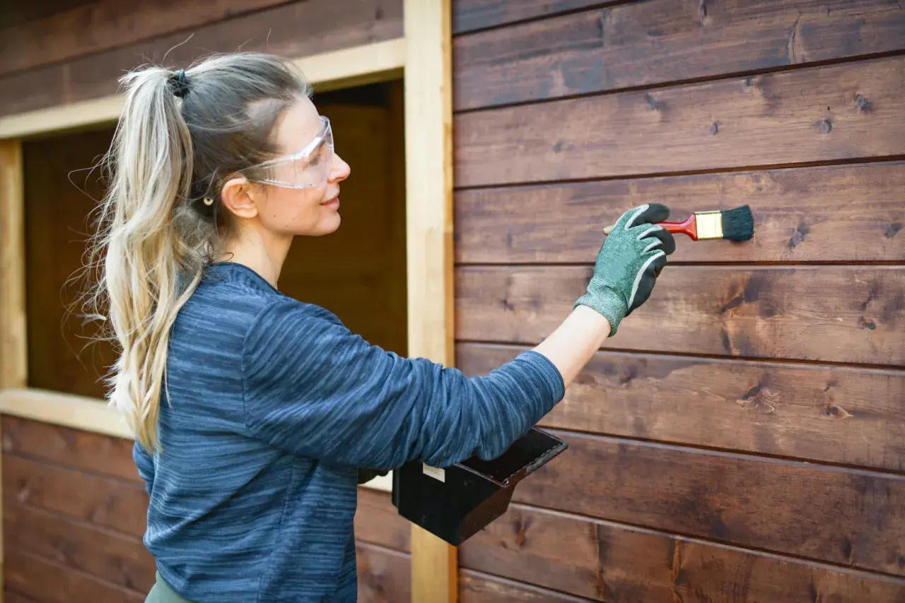 Donna con occhiali protettivi applica vernice su una parete di legno scuro. Si può verniciare sopra l'impregnante per un nuovo look.