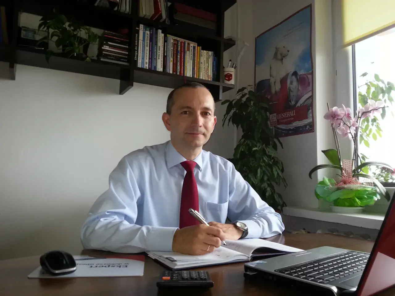Mężczyzna w koszuli i krawacie siedzi przy biurku, pisząc w notatniku. Na biurku laptop, kalkulator i myszka.
