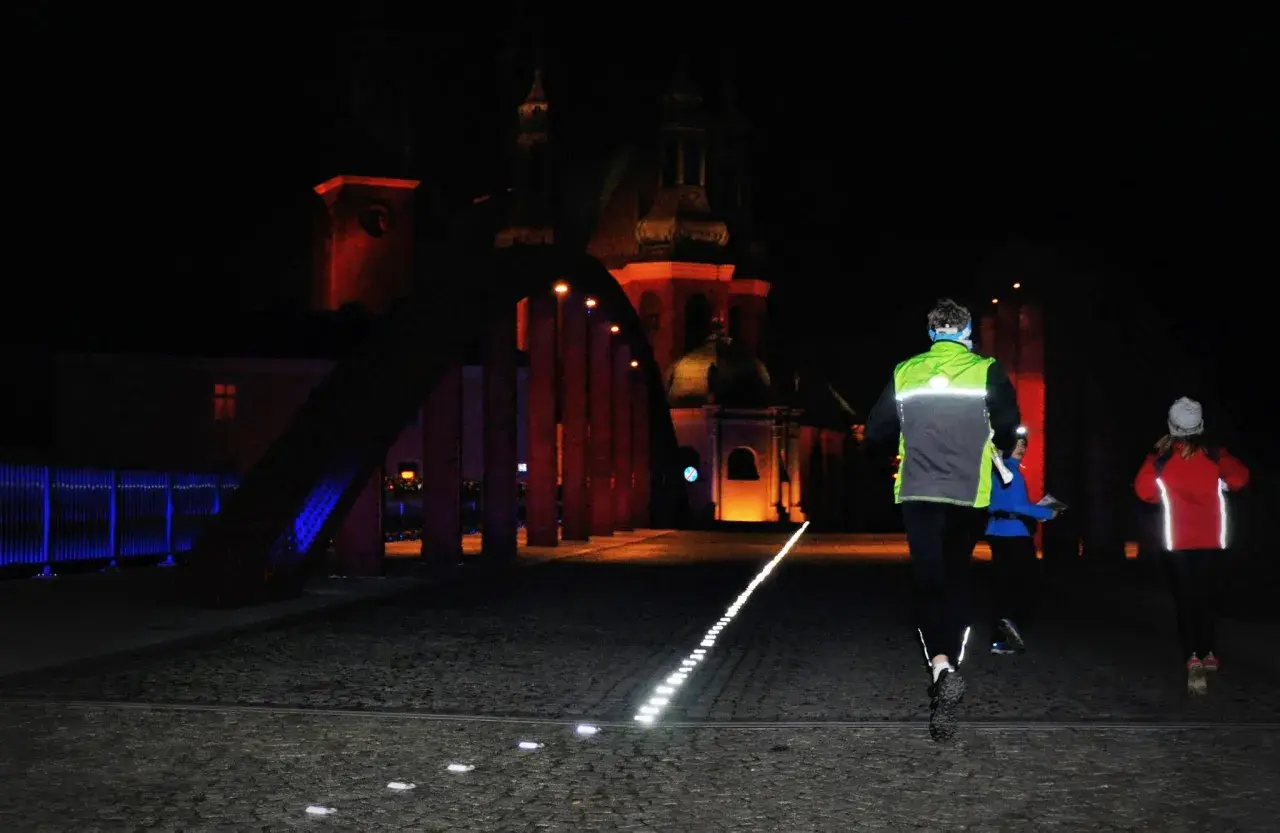 Trzy osoby uprawiające bieganie nocą po brukowanej ulicy, oświetlonej rzędem lamp. W tle widać podświetlony budynek z kopułą.