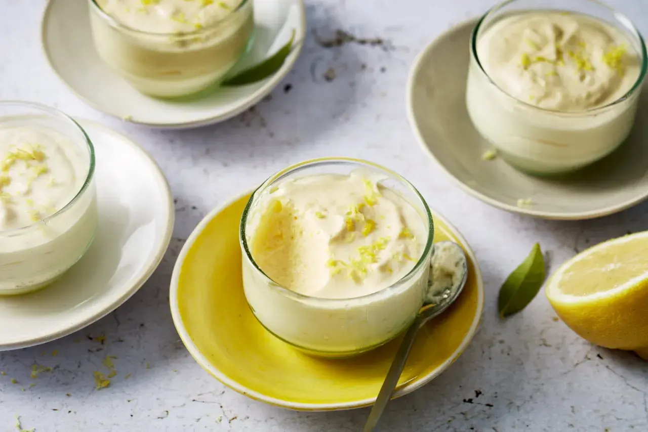 Plusieurs verrines de mousse au citron, garnies de zestes, sur des assiettes claires et jaunes. Un citron coupé est visible à droite.