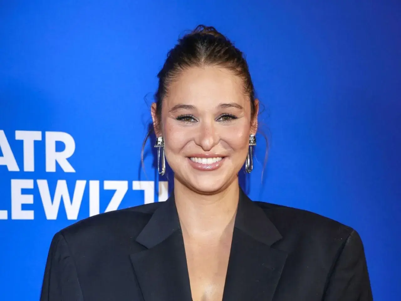 Uśmiechnięta Anna Szymańczyk w czarnej marynarce, z długimi kolczykami. Może właśnie wraca do męża po premierze?