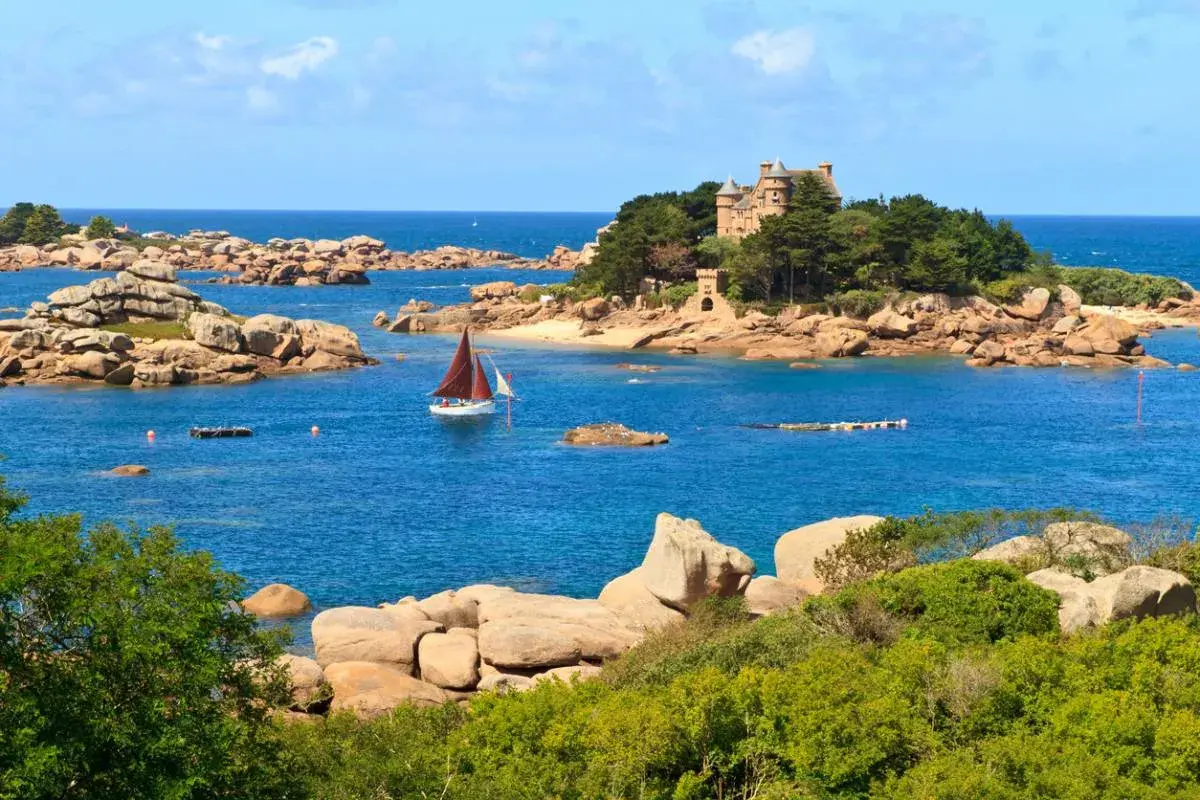 Un voilier navigue près d'un château sur une île rocheuse en Bretagne. Le paysage est typique de la côte bretonne.