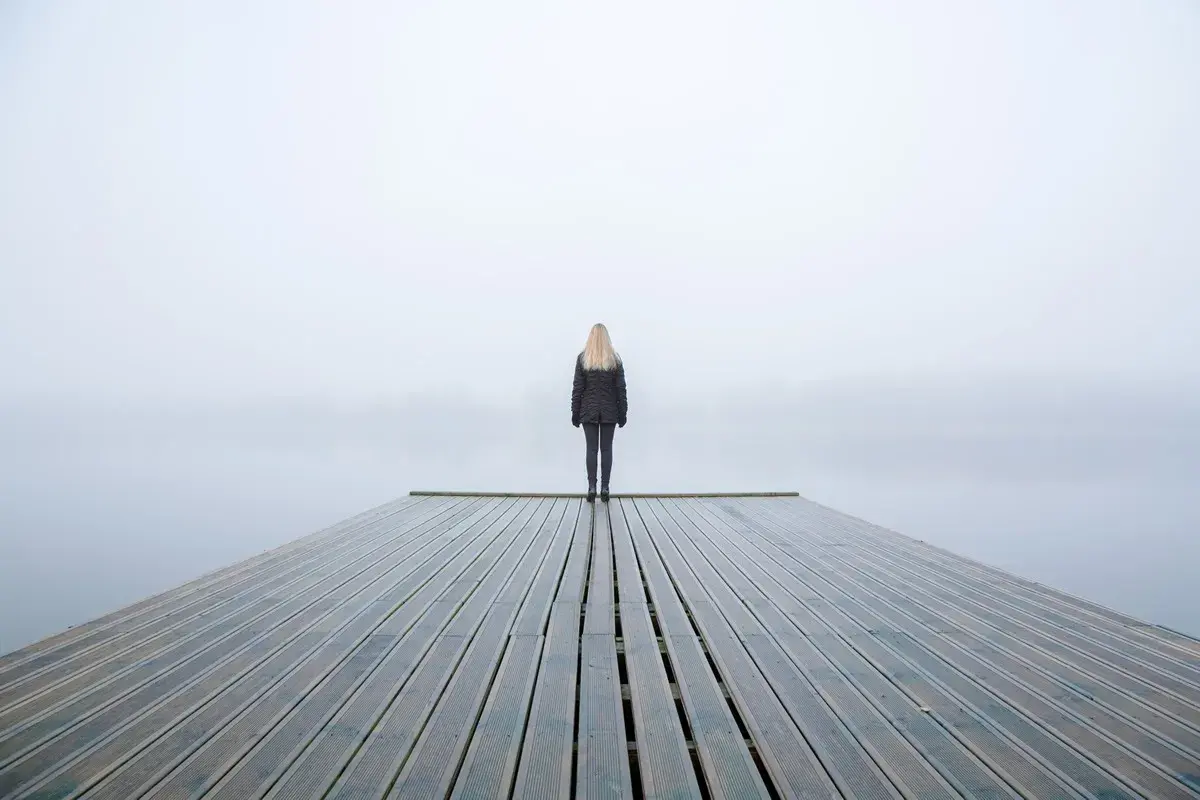 Una persona fredda, con la psicologia che la isola, sta in piedi su un molo di legno in una giornata nebbiosa.