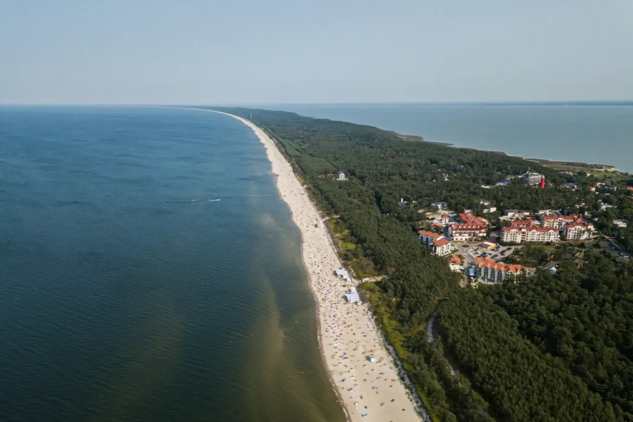 Widok z lotu ptaka na plażę w Krynicy Morskiej, przypominającą wielbłądzi garb. Widać tłumy plażowiczów, las i zabudowania.