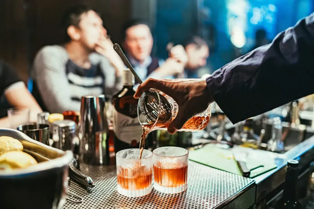 Un barman prépare un cocktail de base, versant un liquide ambré dans deux verres.
