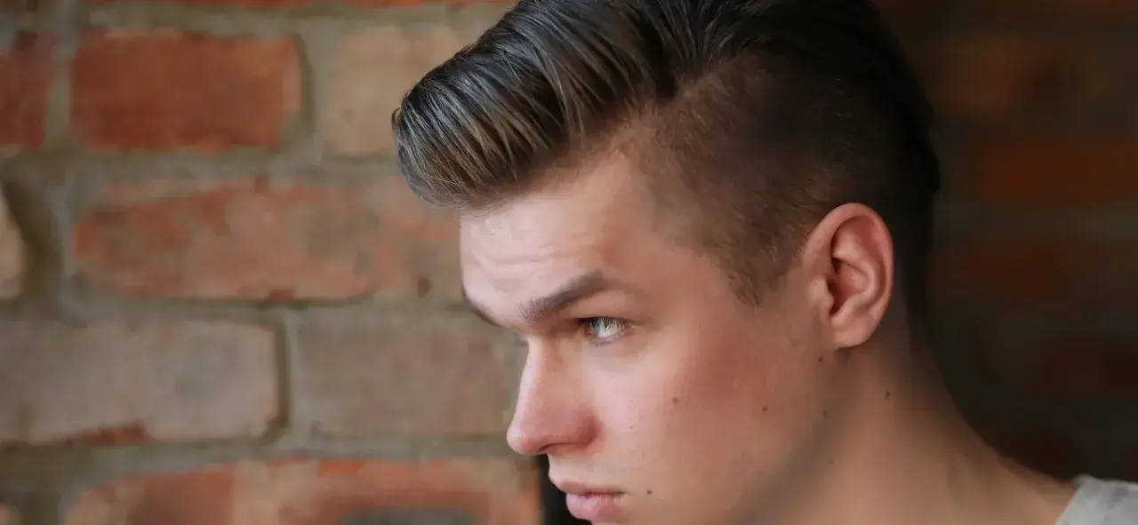 Joven con un corte de pelo moderno, un mid fade en pico, con el cabello peinado hacia arriba y a un lado, frente a una pared de ladrillos.