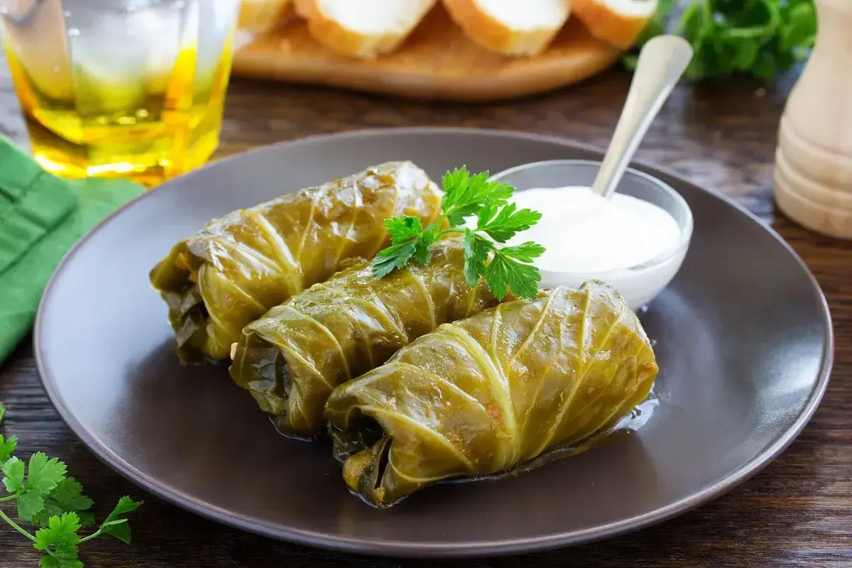 Tre involtini di cavolo cappuccio, guarniti con prezzemolo, serviti con una ciotolina di salsa bianca e un bicchiere di bevanda dorata.