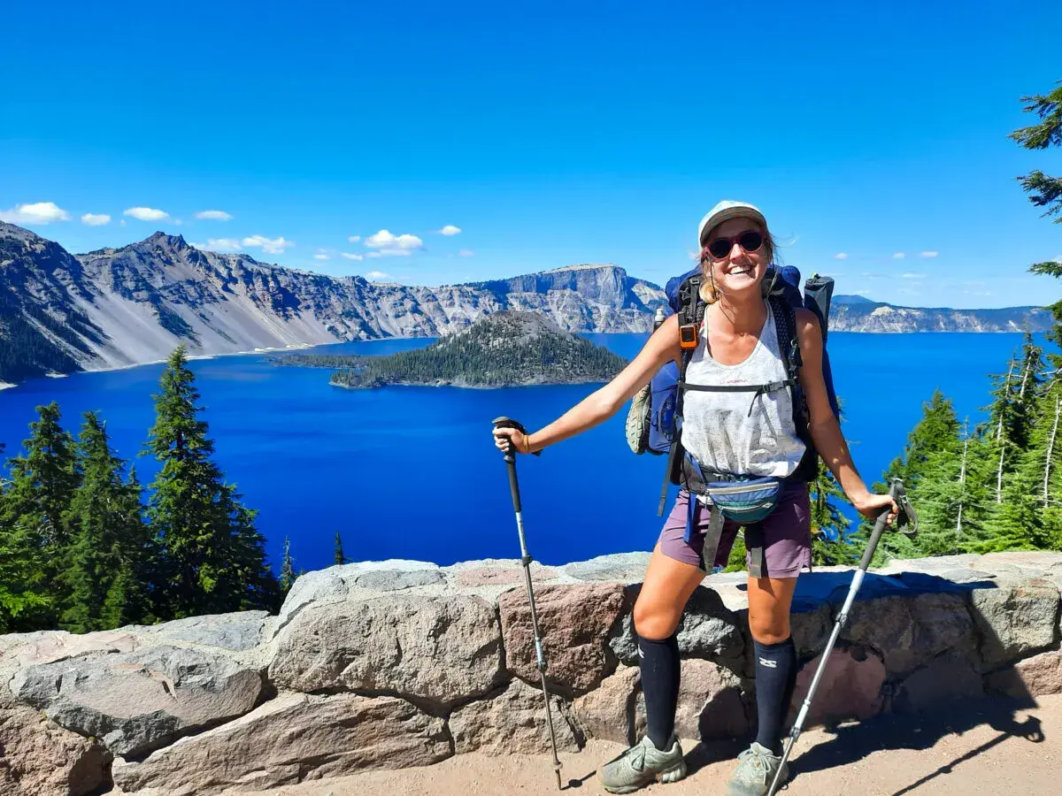 Uśmiechnięta turystka z plecakiem i kijkami, podziwiająca błękitne jezioro. To widok z trasy Pacific Crest Trail, pokazujący, ile się idzie przez takie cuda natury.