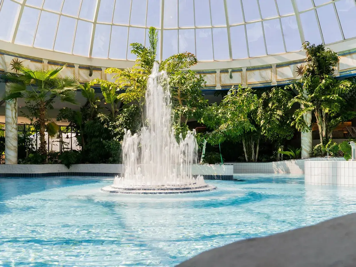 Ein tropischer Poolbereich mit einem sprudelnden Brunnen und Palmen unter einem Glasdach. Perfekt für einen entspannten Urlaub in Hessen.