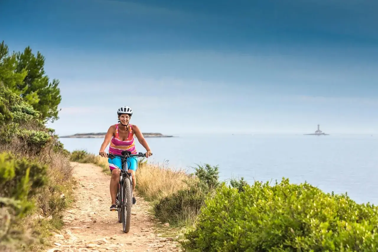 Woman enjoys cycling tours in Slovenia, biking along a coastal path with the sea and a distant lighthouse.