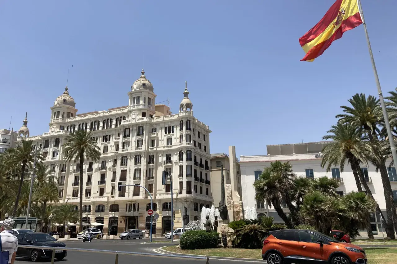 Alicante, pogoda marzec: słoneczny dzień, palmy i hiszpańska flaga nad białym budynkiem.