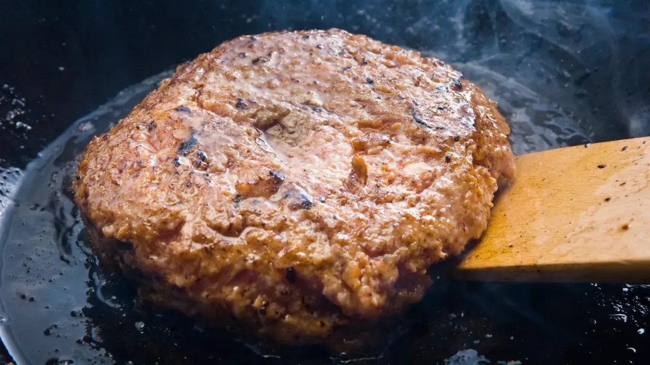 Jugosa carne dorándose en sartén, parte de como hacer hamburguesas caseras. Humo y burbujas de grasa.