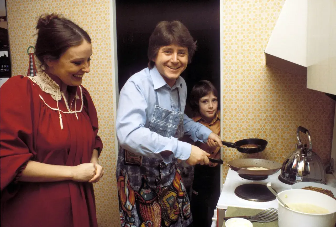 Familie in der Küche, der Vater brät Pfannkuchen nach DDR Rezepten. Ein Kind schaut zu, die Mutter lächelt.