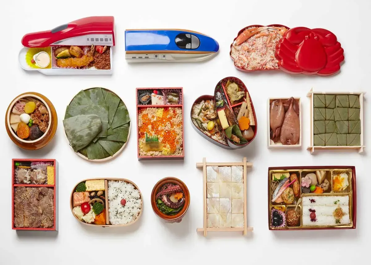 A variety of Japanese bento boxes are displayed, including train-shaped ones, a heart-shaped bento, and those filled with seafood, rice, and other delicacies.