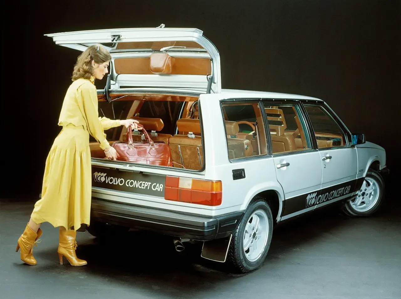 En kvinna i gul klänning lastar en väska i en silverfärgad Volvo concept car från 80-talet.