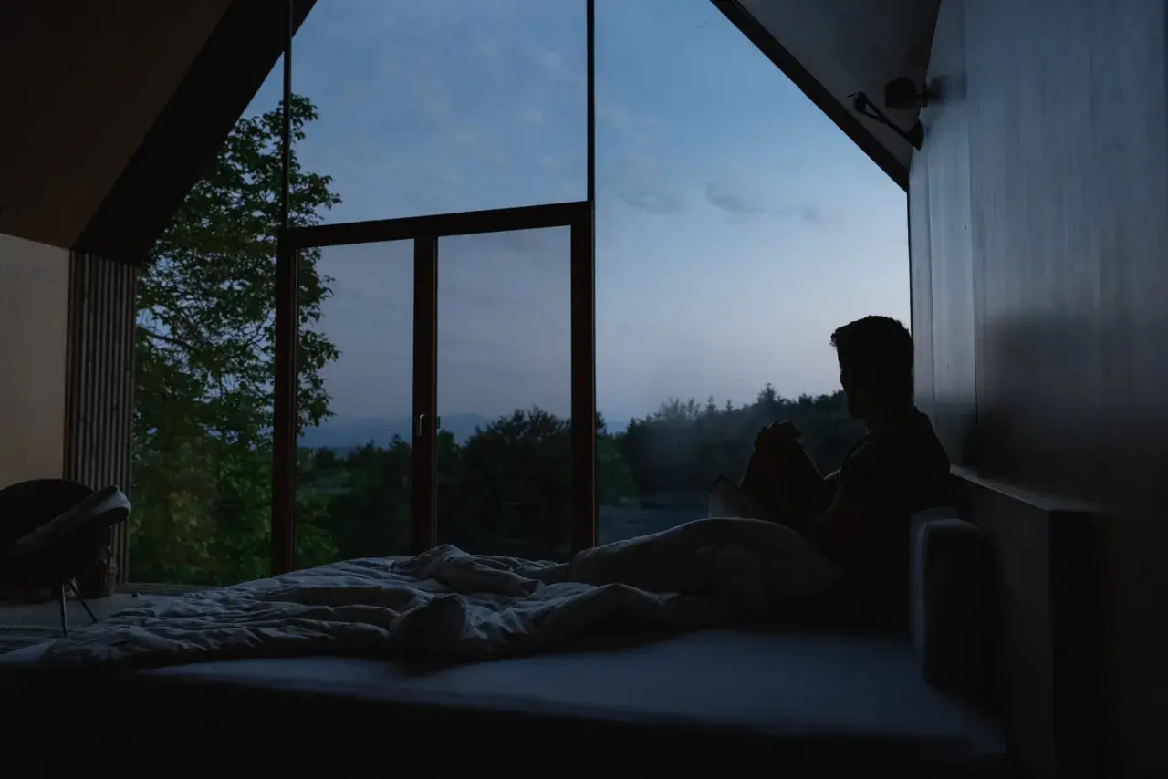Mann sitzt im Bett, blickt aus dem Fenster in die Dämmerung. Ein ruhiger Moment, der den schlaf wach rhythmus widerspiegelt.