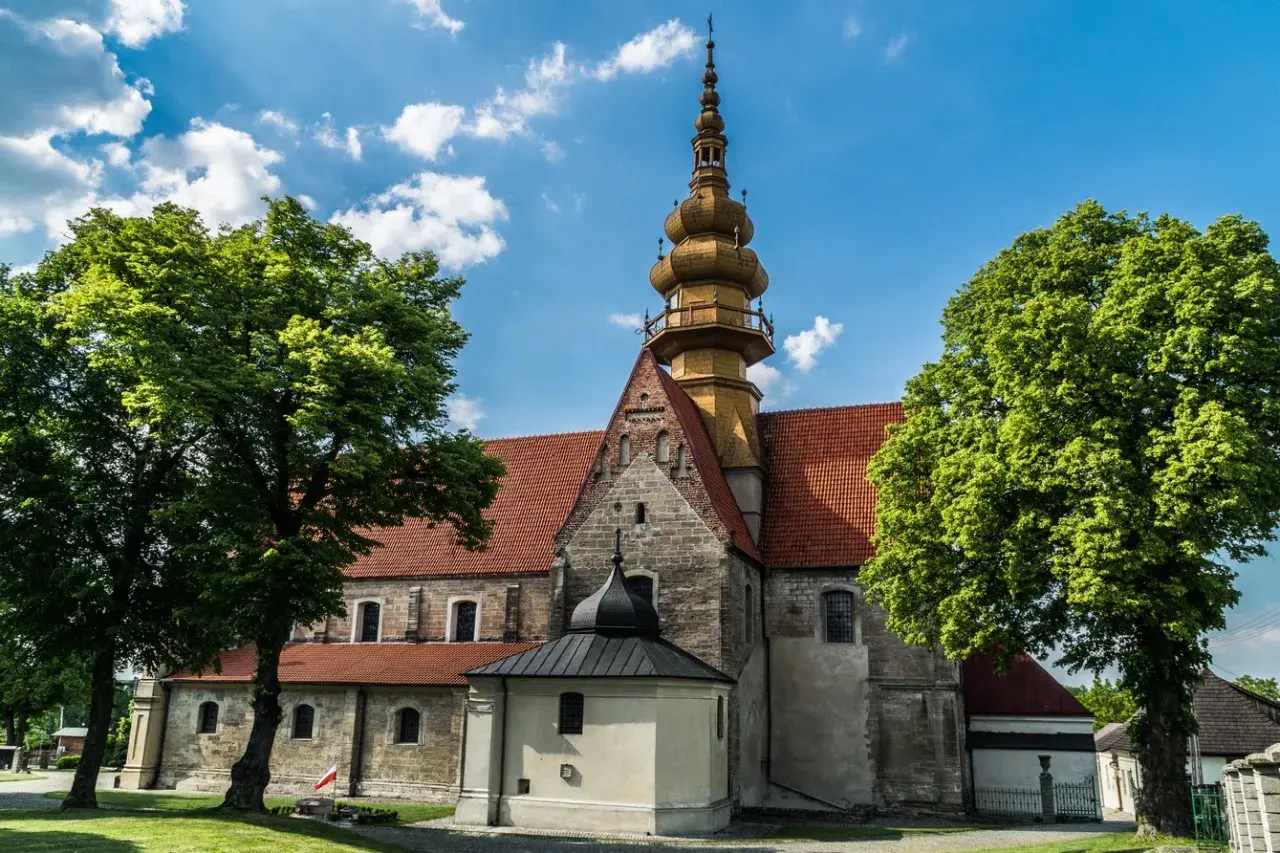 Kamienna świątynia z żółtą wieżą i czerwonym dachem, otoczona zielenią. W oddali widać fragmenty zabudowań, przypominające krajobraz wokół **zamku Krzyżtopór**.