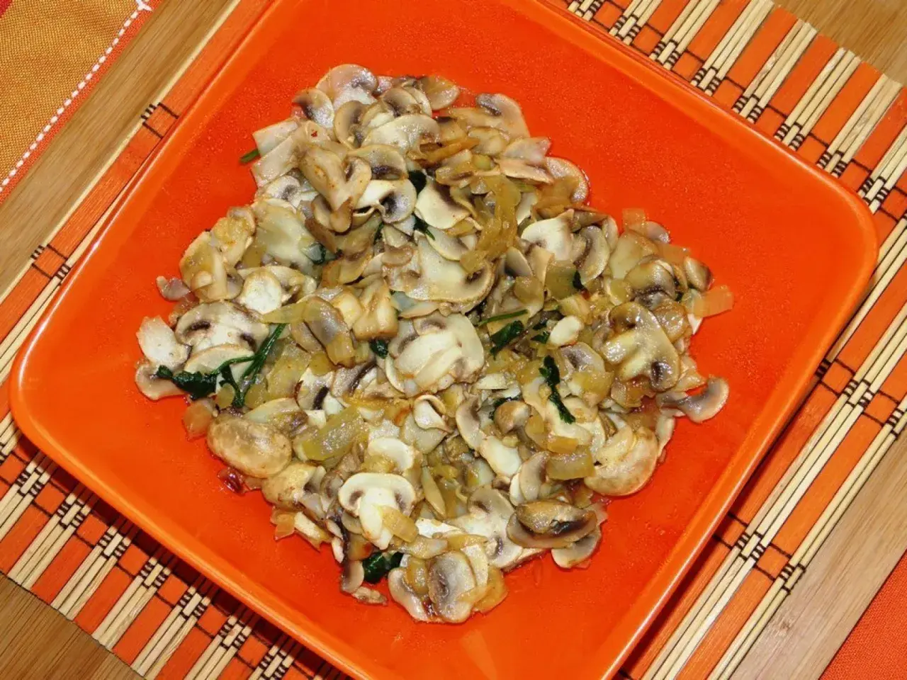 Champiñones salteados con cebolla y espinacas, servidos en un plato naranja sobre un mantel de bambú.
