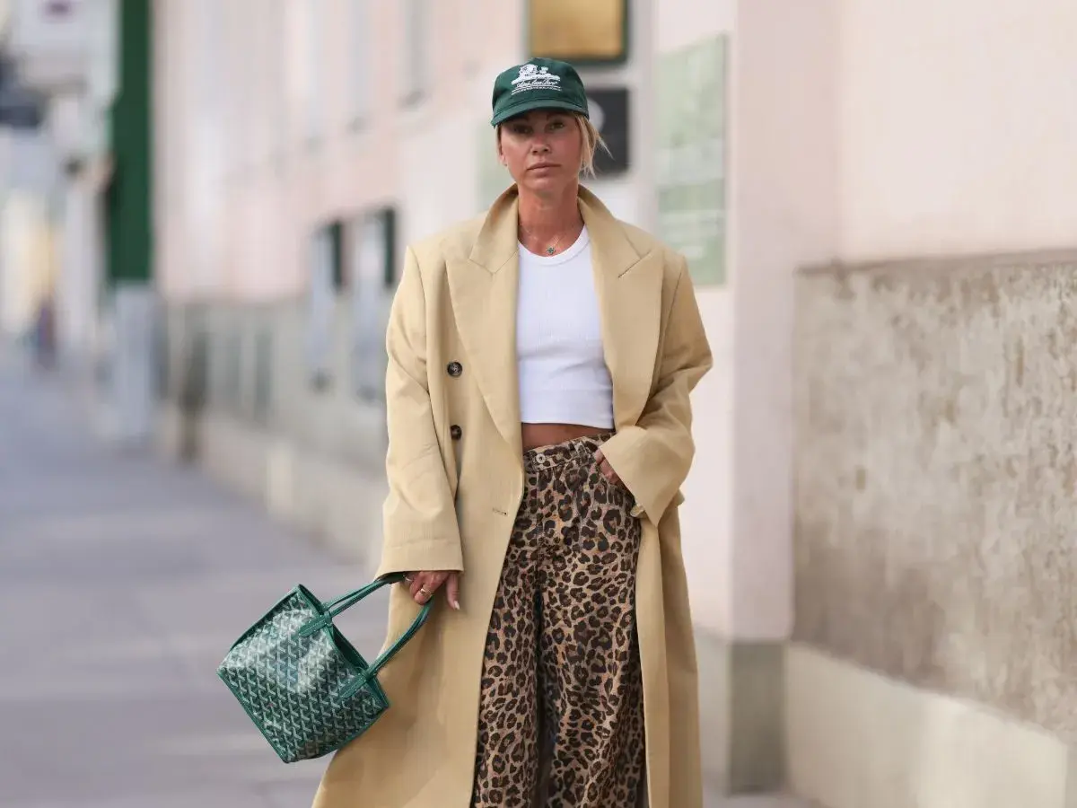 Modna kobieta w beżowym płaszczu, białym topie i spodniach w panterkę. Zielona torebka i czapka dopełniają stylizację.