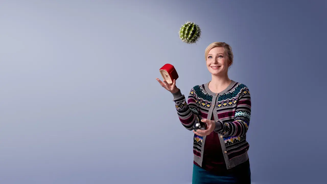 Kvinna jonglerar med en kaktus, en röd väckarklocka och en metallkula. Hon ser glad ut, som om hon klarar allt. Osa 2015.
