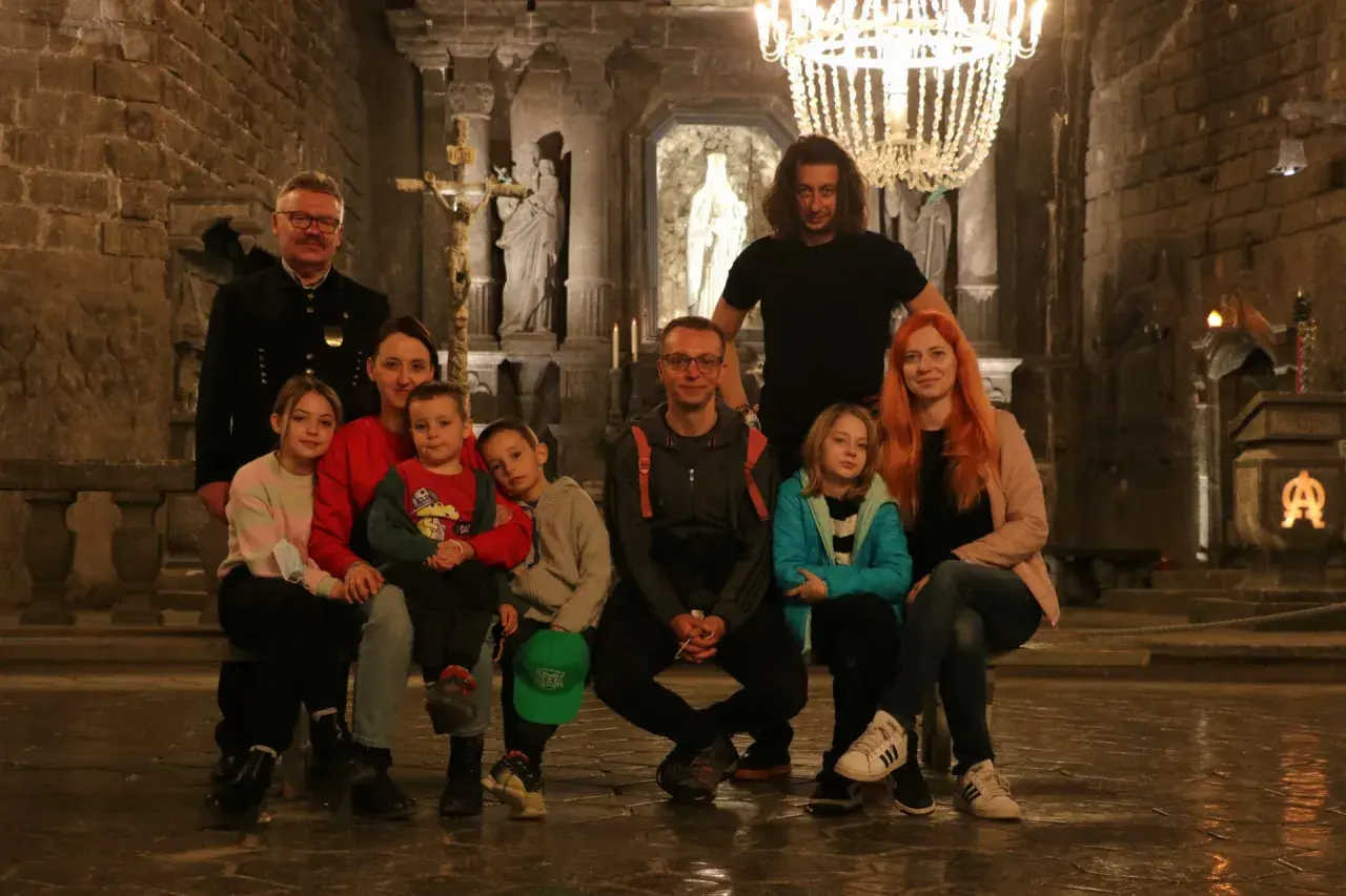 Rodzina zwiedza kopalnię soli w Wieliczce. Dzieci z rodzicami siedzą na ławce w podziemnej kaplicy, podziwiając solne rzeźby i żyrandol.