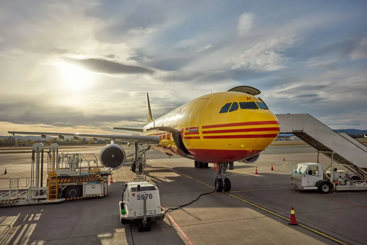 Żółty samolot DHL z czerwonymi pasami na płycie lotniska, gotowy do lotu do USA.