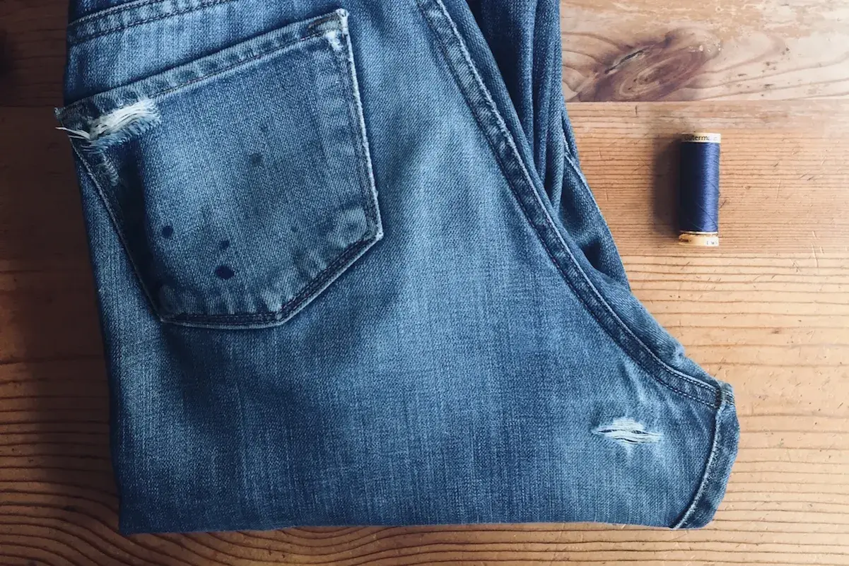 A pair of blue jeans with a small rip on the back pocket and a spool of navy thread, ready for how to patch jeans.