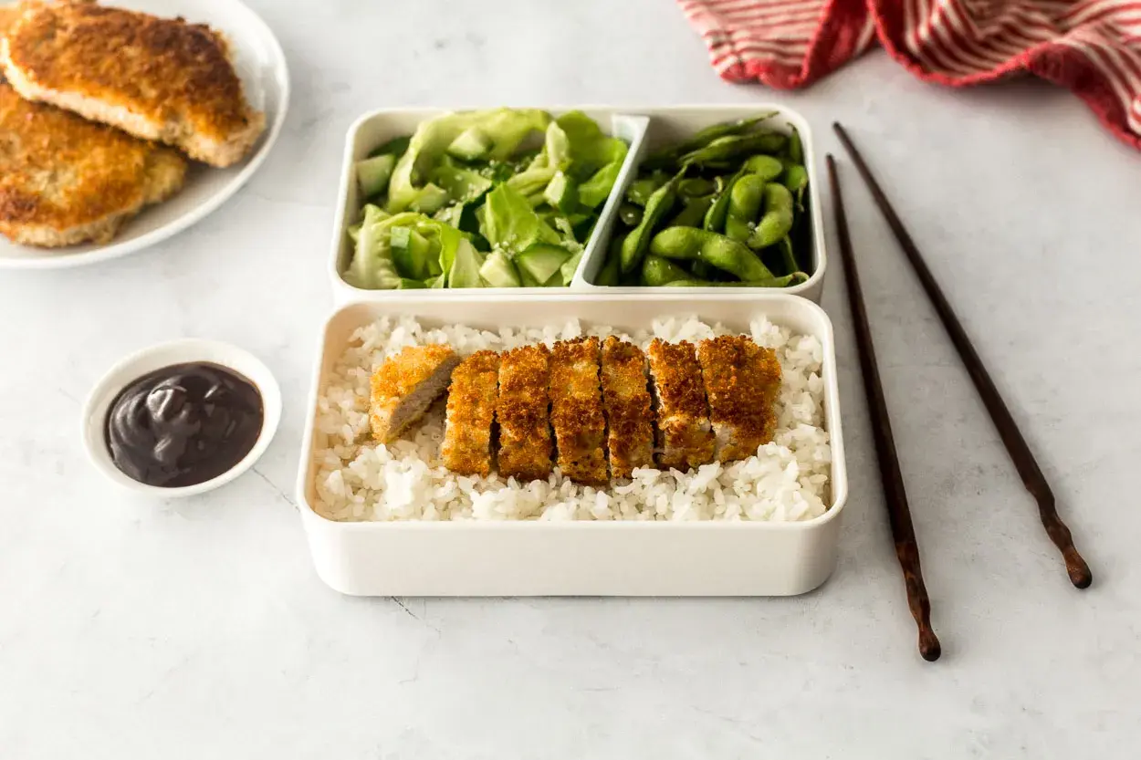 A delicious pork katsu bento box with rice, salad, edamame, and sauce.