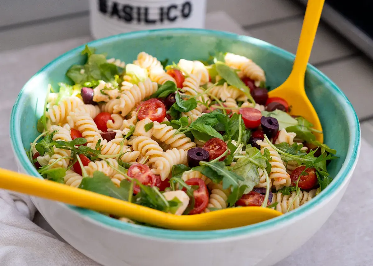 Ensalada de pasta espectacular con fusilli, tomates cherry, aceitunas negras y rúcula, servida en un cuenco azul con cucharas amarillas.