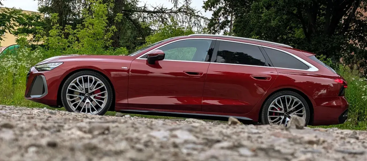 Lśniące, czerwone Audi A6 C9 Avant stoi na kamienistym podłożu, otoczone zielenią.