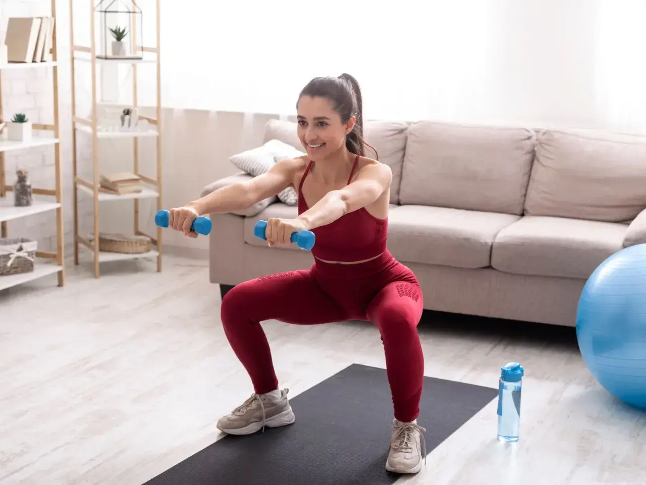 Uśmiechnięta kobieta wykonuje przysiady z hantlami, ćwicząc w domu. Intensywny trening tabata w domowym zaciszu.