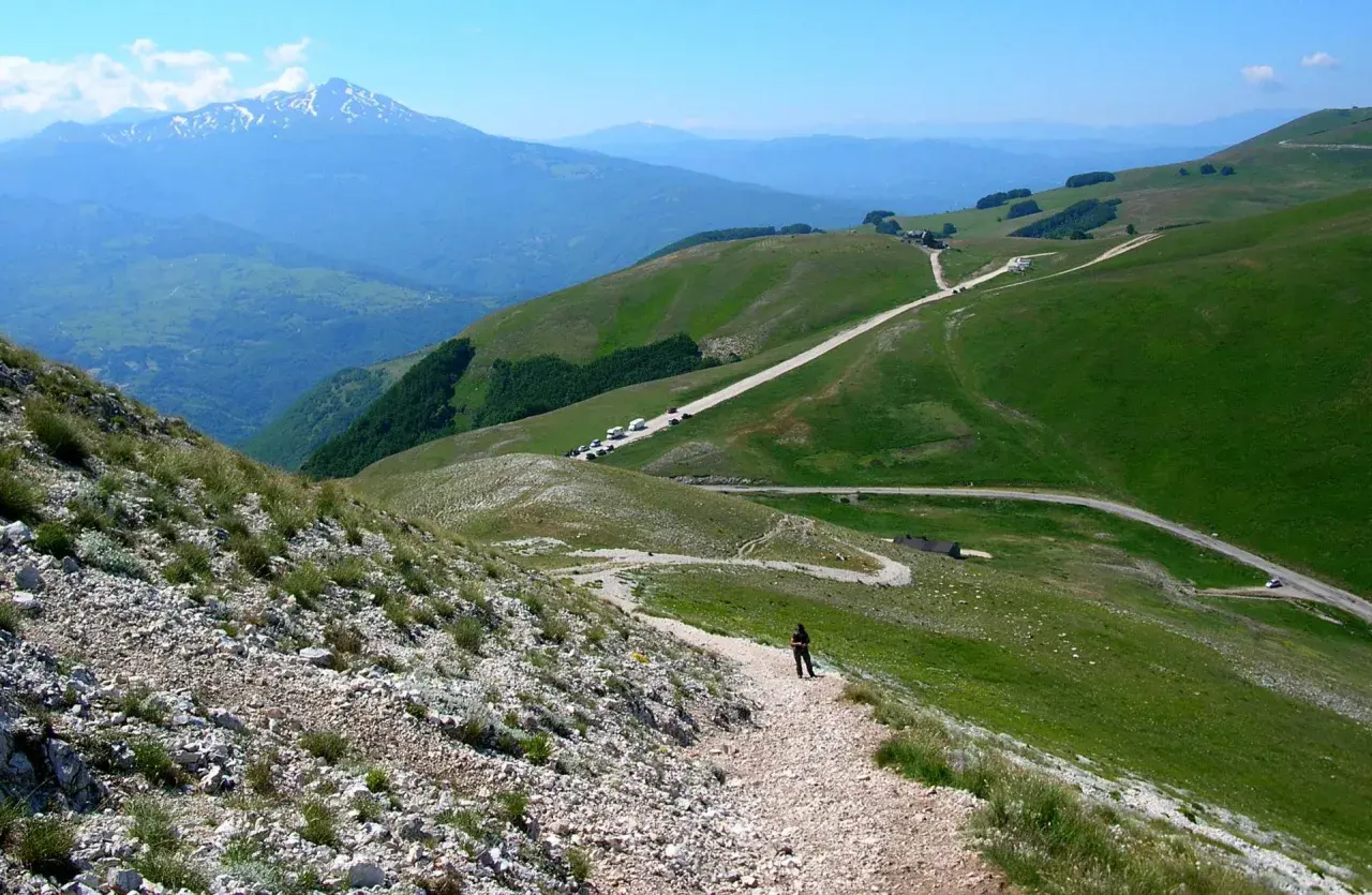 A person walks on a gravel path on a green mountain slope with a road winding through the hills and a snow-capped mountain in the distance.