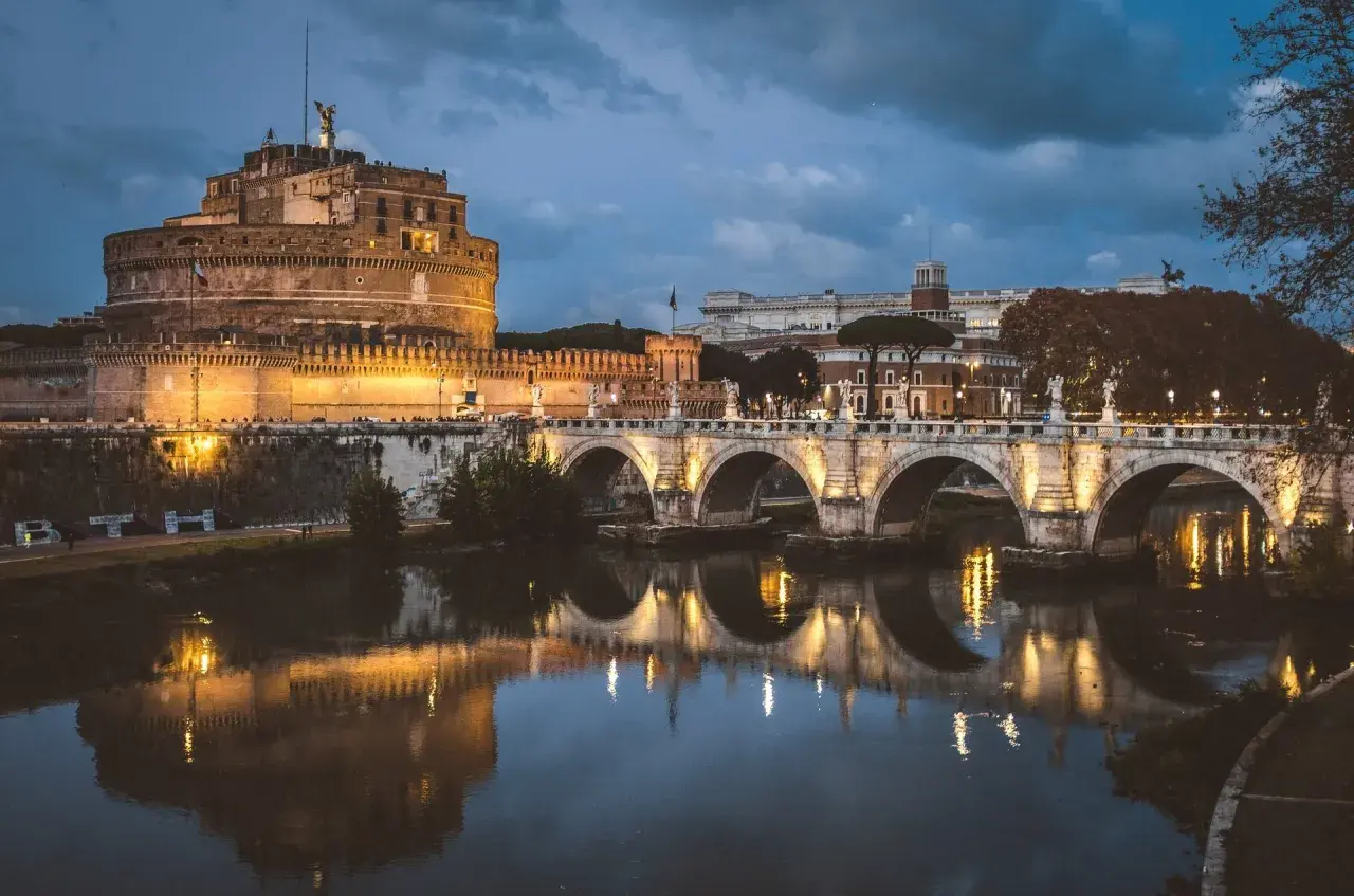 Le Castel Sant'Angelo, illuminé la nuit, se reflète dans le Tibre, avec un pont arqué en avant-plan.