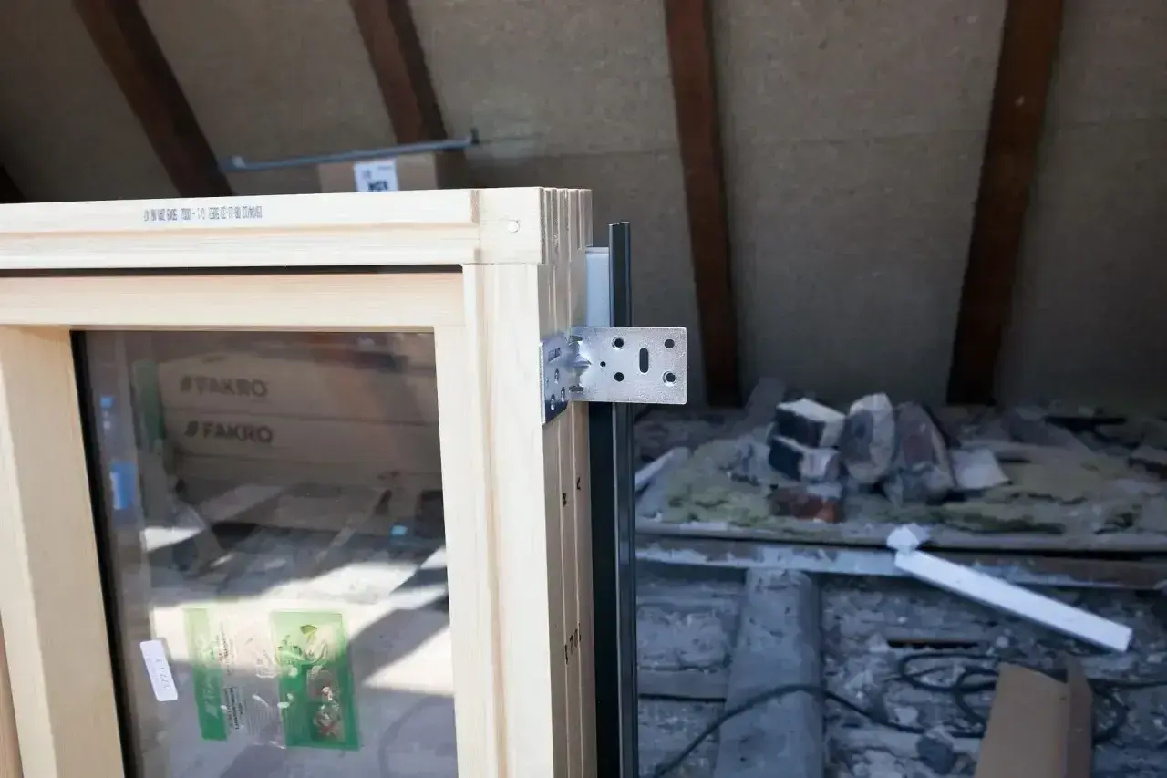 Drewniane okno dachowe z metalowym kątownikiem, gotowe do montażu. W tle widać materiały budowlane i elementy konstrukcji poddasza.