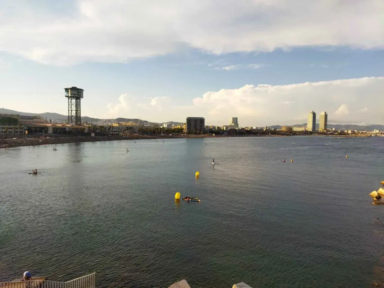 Vista de la playa y el mar con el icónico hotel vela al fondo. Personas disfrutan del agua en tablas de paddle surf.