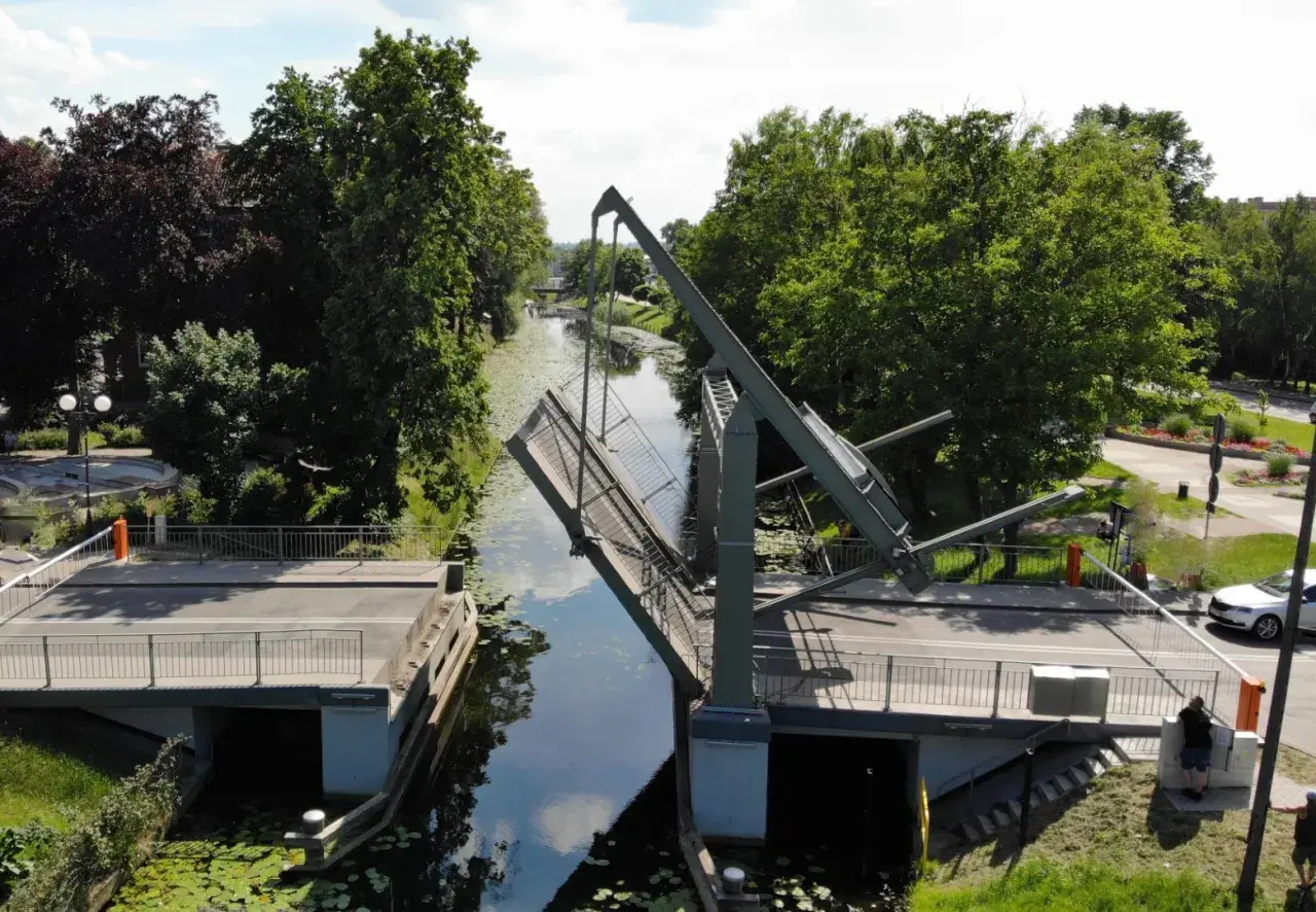 Podniesiony most zwodzony w Nowym Dworze Gdańskim, nad kanałem otoczonym zielenią.