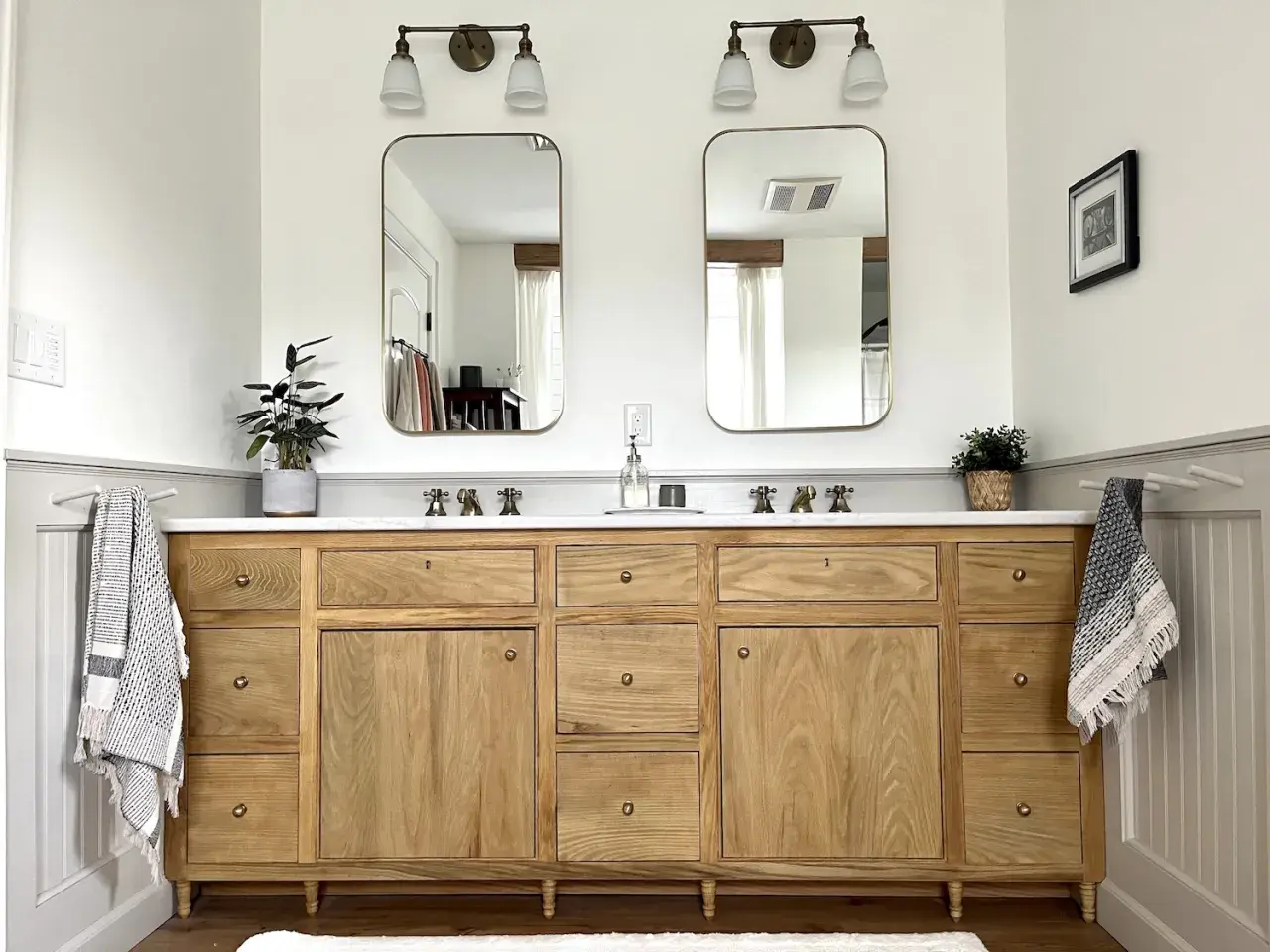 A beautiful DIY bathroom vanity with a natural wood finish, double sinks, and brass fixtures. Two framed mirrors hang above, reflecting the bright, airy space.