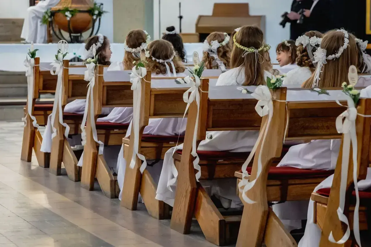 Dzieci w bieli, z wiankami na głowach, siedzą w ławkach kościelnych. Czekają na ważny moment, zastanawiając się, ile trwa komunia.
