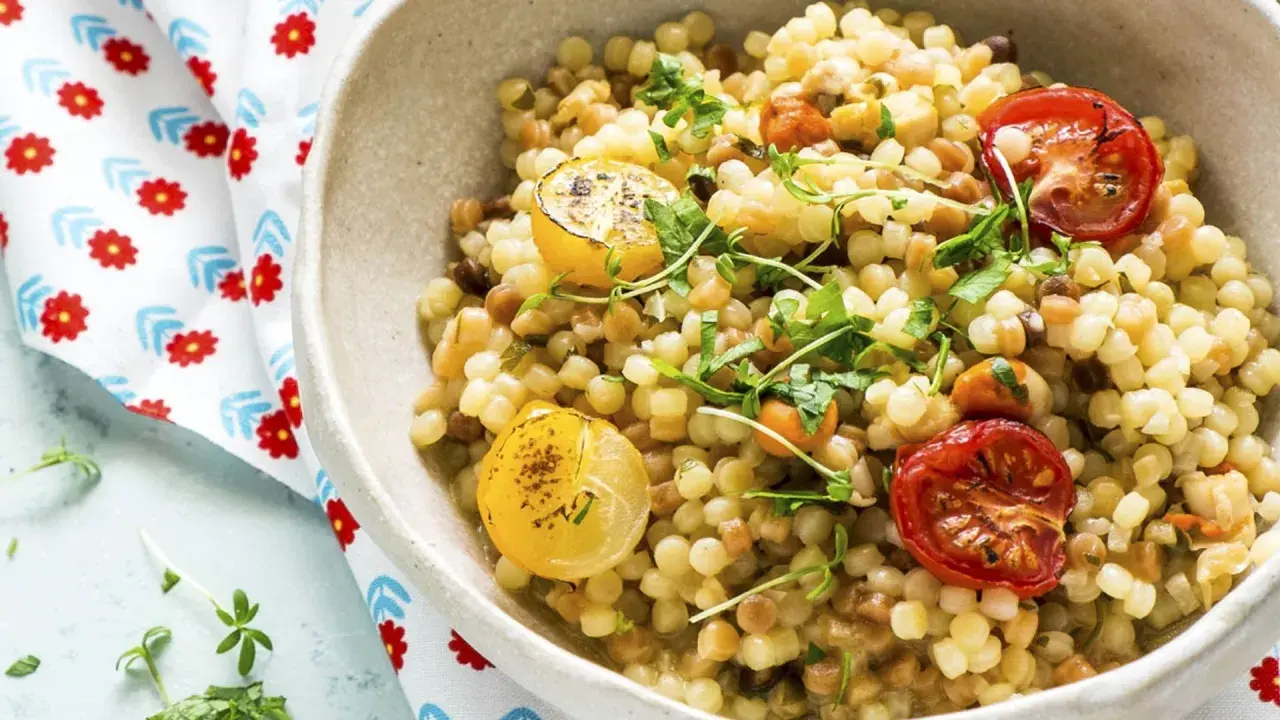 Un plato de pasta pequeña, que es semola, con tomates cherry asados y rodajas de limón, adornado con hierbas frescas.