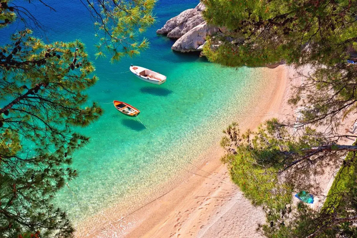 Idealne tanie wczasy Chorwacja z wyżywieniem: dwie łódki kołyszą się na turkusowej wodzie przy piaszczystej plaży, otoczonej sosnami.