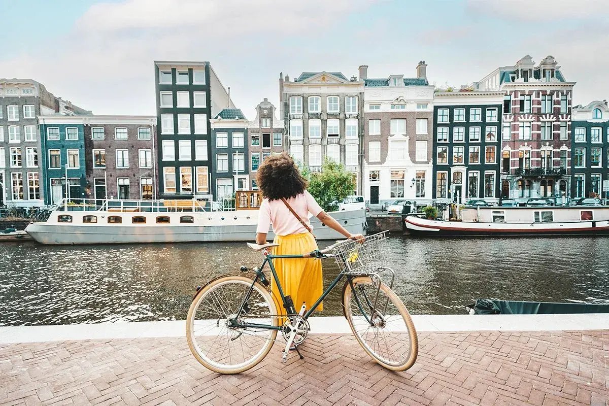 Kobieta z rowerem na tle amsterdamskich kamienic i kanału. Idealne miejsce na odkrywanie tras rowerowych Europa.